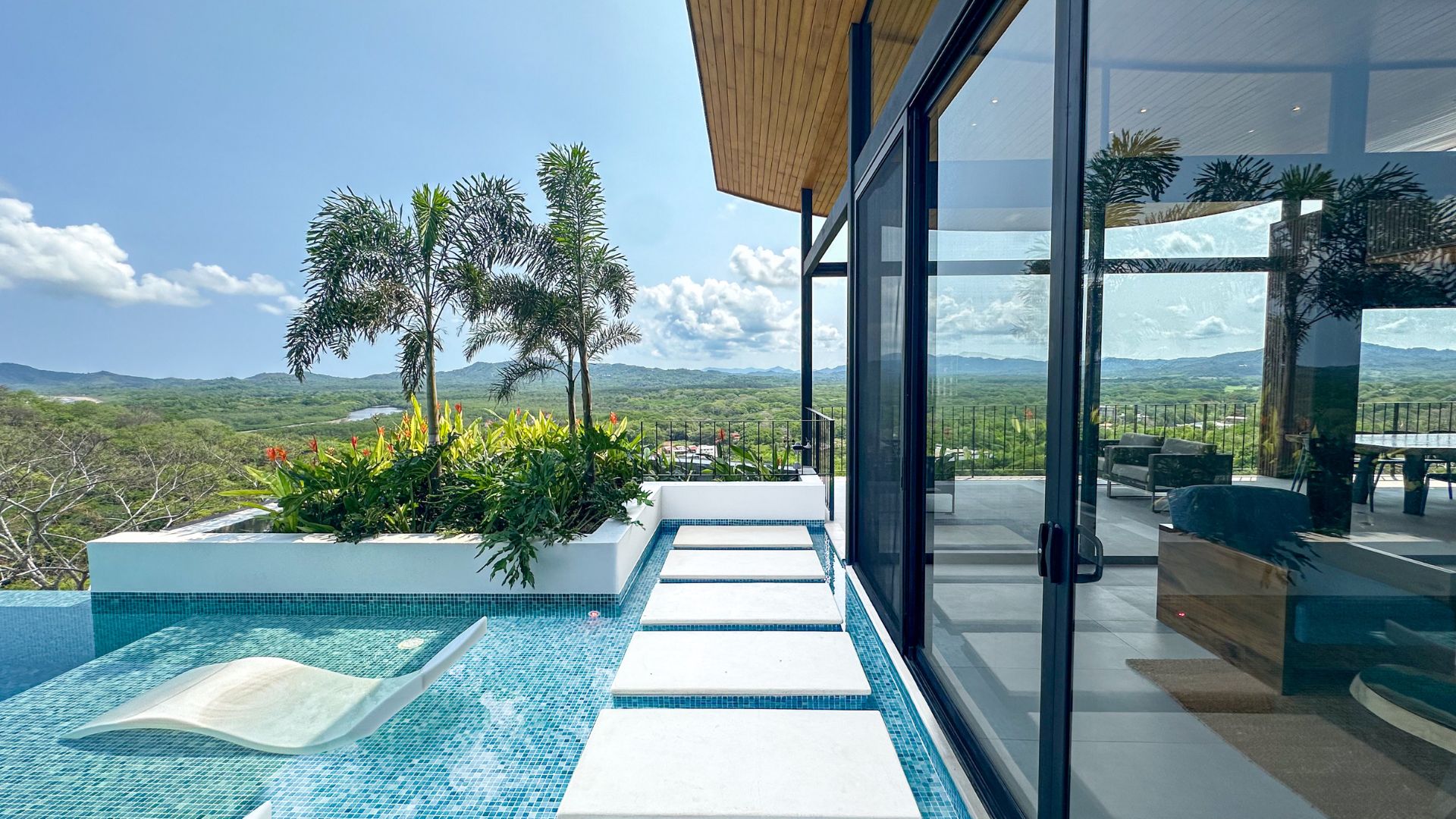 Modern house with large glass windows overlooks a lush green landscape. A pool with stepping stones and a lounge chair is in the foreground, surrounded by tropical plants.