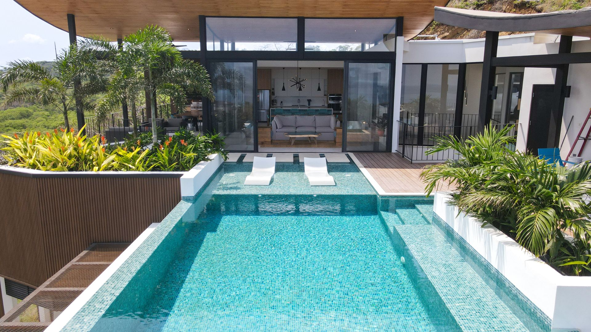 Modern house with floor-to-ceiling windows and outdoor pool, featuring two white lounge chairs submerged in shallow water, surrounded by tropical plants.