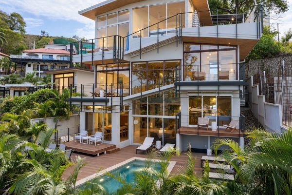 Modern multi-story house with large glass windows, multiple balconies, outdoor seating areas, and a swimming pool, surrounded by tropical plants—this is Casa Uno Mas, where contemporary design meets tropical elegance.